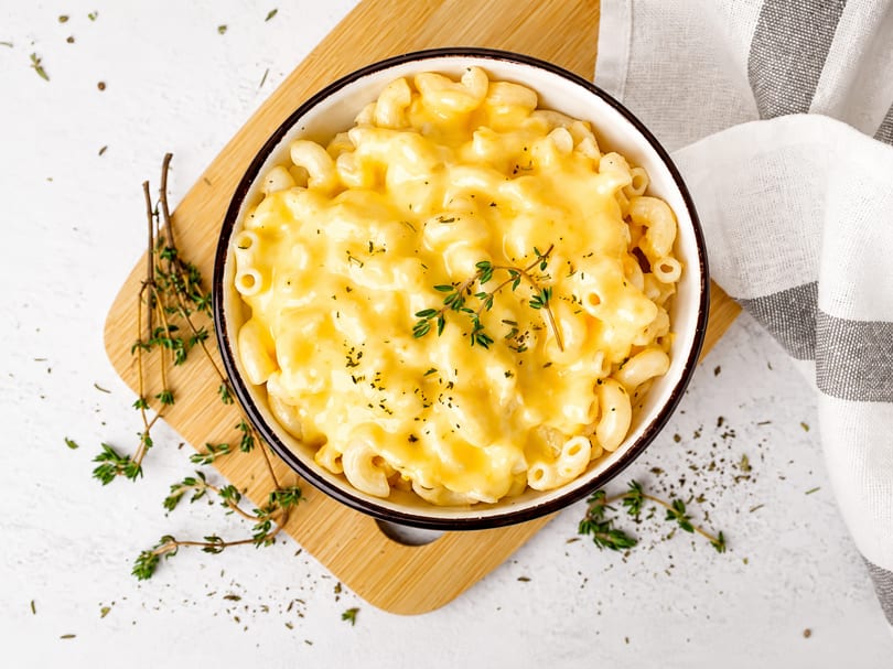 Creamy macaroni and cheese in a white bowl, garnished with fresh thyme, on a wooden board.