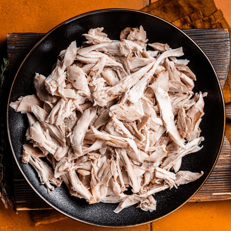 Shredded chicken in a black bowl, on a wooden board with an orange background.